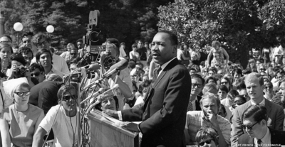 Image shows Dr. Martin Luther King Jr. standing at a podium with microphones, surrounded by a large crowd of students.